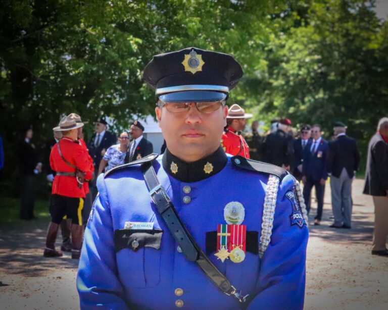 A Truro Police officer, identified by the name tag as Cst. B. Bowden, stands facing the camera in full ceremonial blue uniform adorned with medals and a shoulder cord. He wears a formal cap and glasses. In the background, several RCMP officers in red serge uniforms and attendees in suits and dresses are gathered outdoors under trees, suggesting a formal or commemorative event.