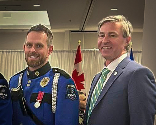A Truro Police officer in full blue ceremonial dress uniform stands beside Nova Scotia Premier Tim Houston, who is wearing a blue suit and green-striped tie. They are smiling and posing indoors in front of a Canadian flag.