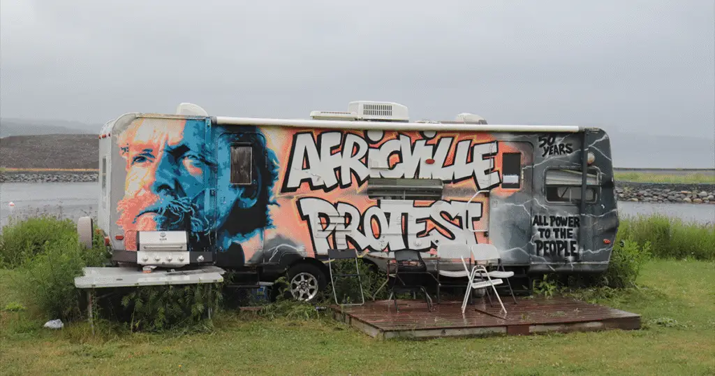 A weathered RV parked near the waterfront is covered in vibrant graffiti art. The left side features a large, stylized portrait of a man with a beard in orange, blue, and black tones. Bold graffiti text across the side reads “AFRICVILLE PROTEST.” Additional text on the right side says “50 YEARS” and “ALL POWER TO THE PEOPLE.” Several empty chairs and a small deck are set up in front of the RV, with overgrown grass surrounding the area under a cloudy sky.