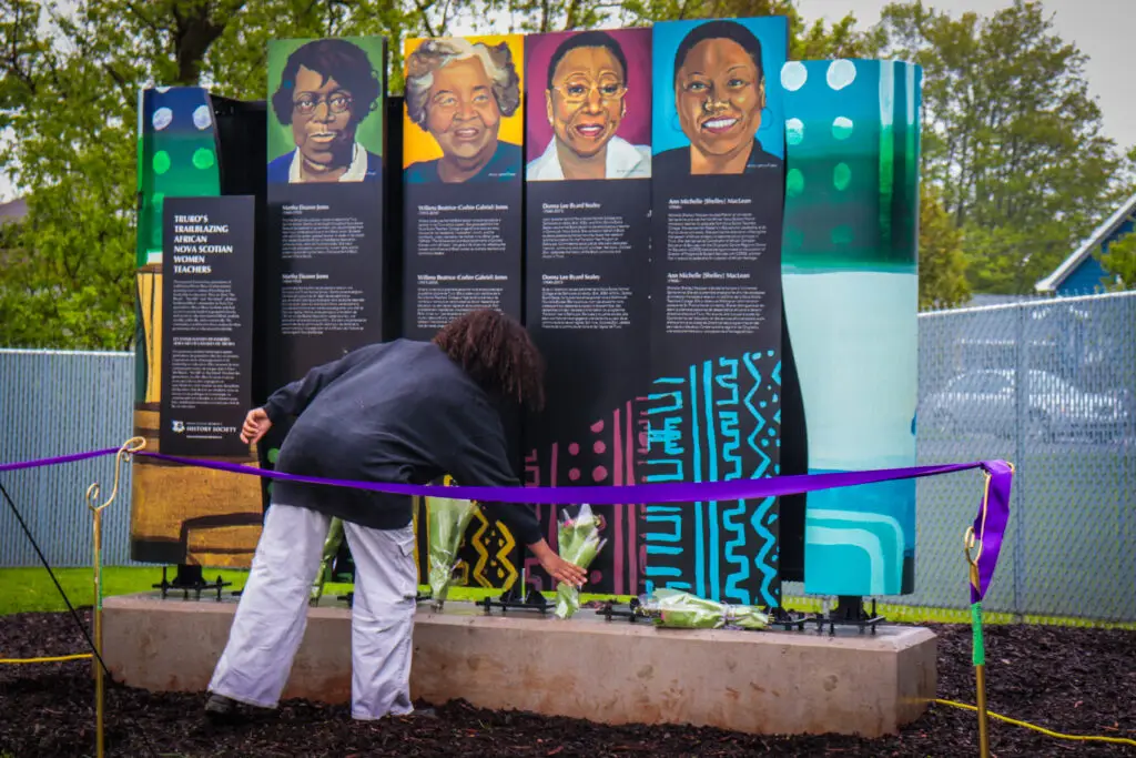 A person places flowers at a public monument featuring painted portraits and biographies of five African Nova Scotian women educators, displayed behind a purple ribbon during a park dedication ceremony in Truro, Nova Scotia.