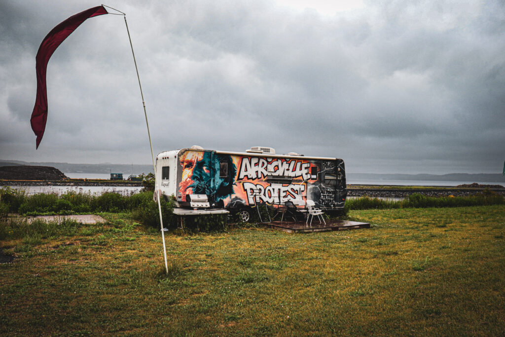 Eddie Carvery’s protest trailer sits on grassy land at the Africville site, painted with the words “Africville Protest,” with a flag flying nearby under an overcast sky.