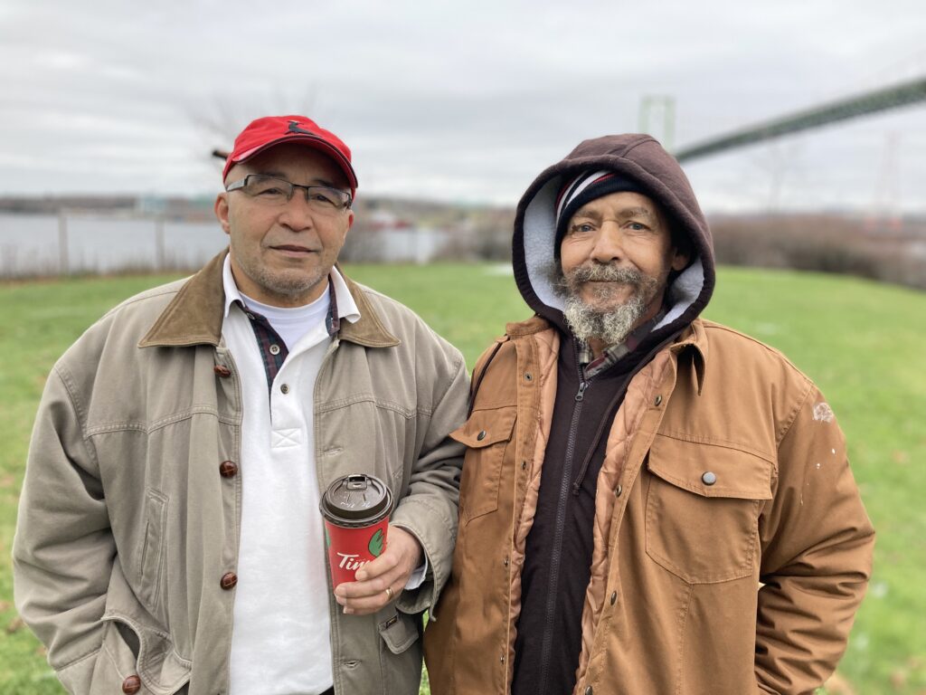 Irvine Carvery and Eddie Carvery stand outdoors at the Africville site, wearing jackets, with the Halifax Harbour and the Macdonald Bridge visible in the background.