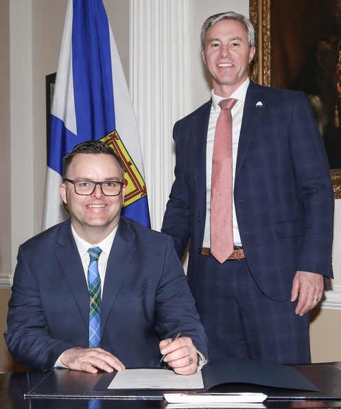 Nova Scotia’s Minister of Education and Early Childhood Development, Brendan Maguire, seated at a desk with Premier Tim Houston standing beside him during a formal signing or announcement at Province House.