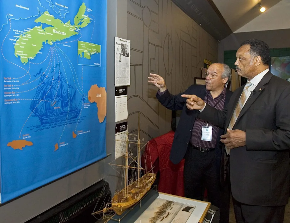 Dr. Leslie “Les” Oliver and Rev. Jesse Jackson stand inside the Black Cultural Centre for Nova Scotia in 2009, gesturing toward a wall display map tracing African Nova Scotian and African diasporic history, with a model ship exhibit in the foreground.