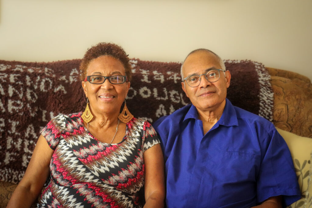 Donna Paris and David Zapparoli seated side by side on a couch, smiling at the camera, with a patterned textile draped behind them.